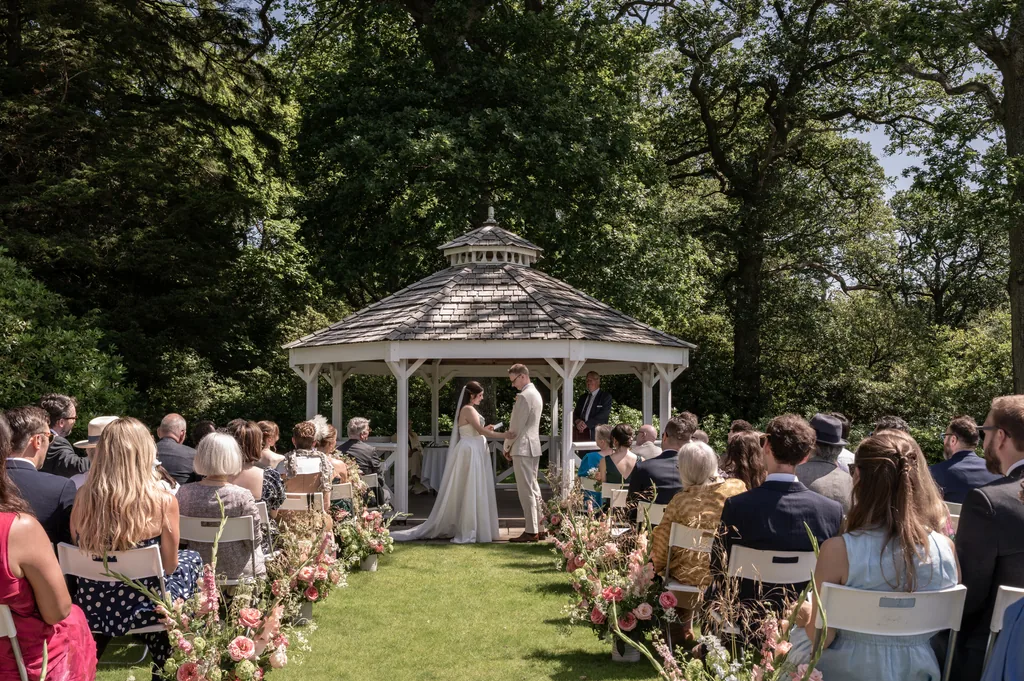 Outdoor wedding ceremony at a white gazebo with guests seated among woodland trees