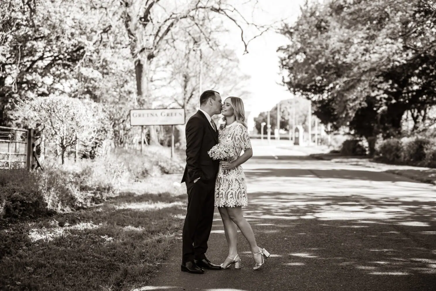 Couple kissing on a tree-lined road beside the Gretna Green sign