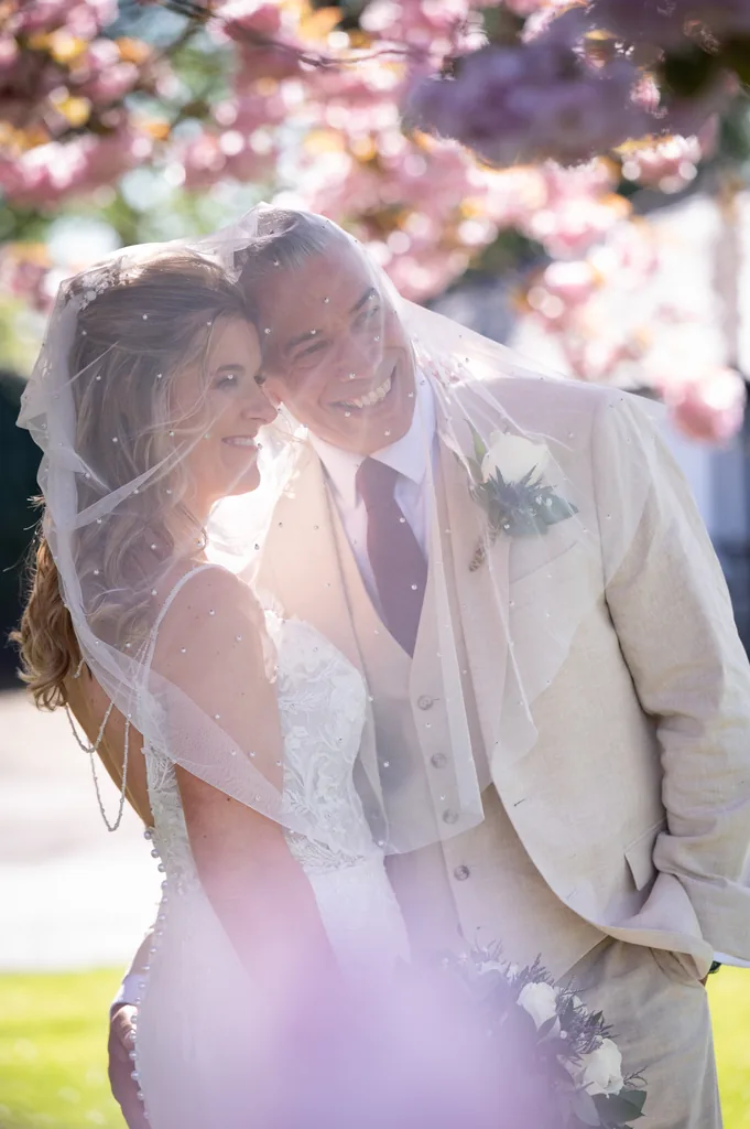 Bride and groom laughing together beneath a bridal veil framed by pink blossom