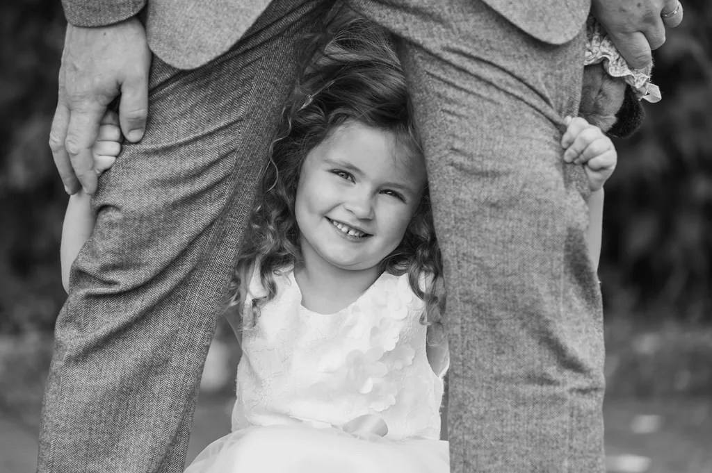 Black-and-white portrait of a smiling flower girl framed between a wedding guest’s legs