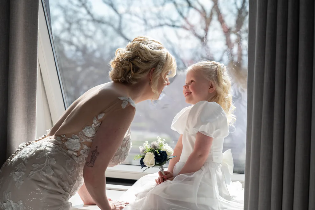 Bride and flower girl leaning towards each other beside a sunlit window