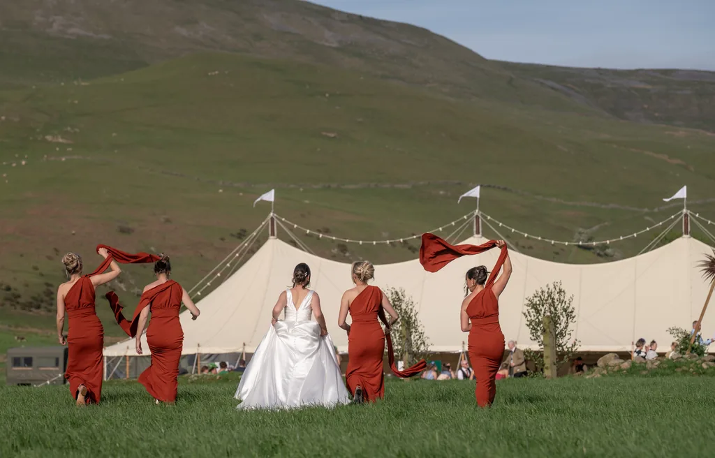 Bride and bridesmaids in rust dresses walking across a grassy hillside towards a marquee