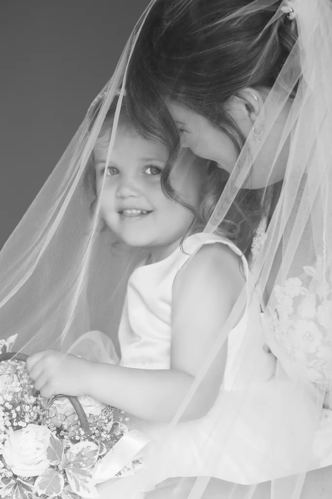 Black-and-white close-up of a bride smiling down at a young flower girl beneath her veil