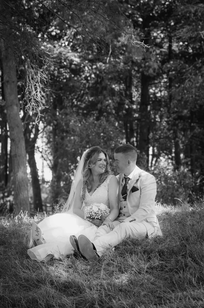 Black-and-white portrait of a bride and groom sitting together on the grass in a forest