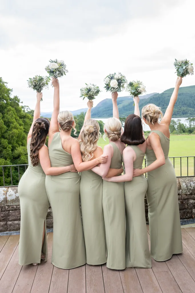 Five bridesmaids in olive-green dresses raising their bouquets toward a mountain backdrop