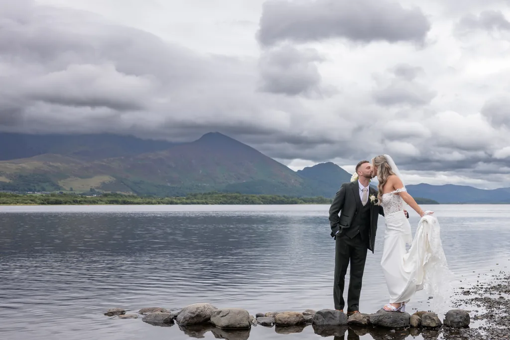 Bride and groom standing on a lakeshore with mountains and dramatic clouds behind them