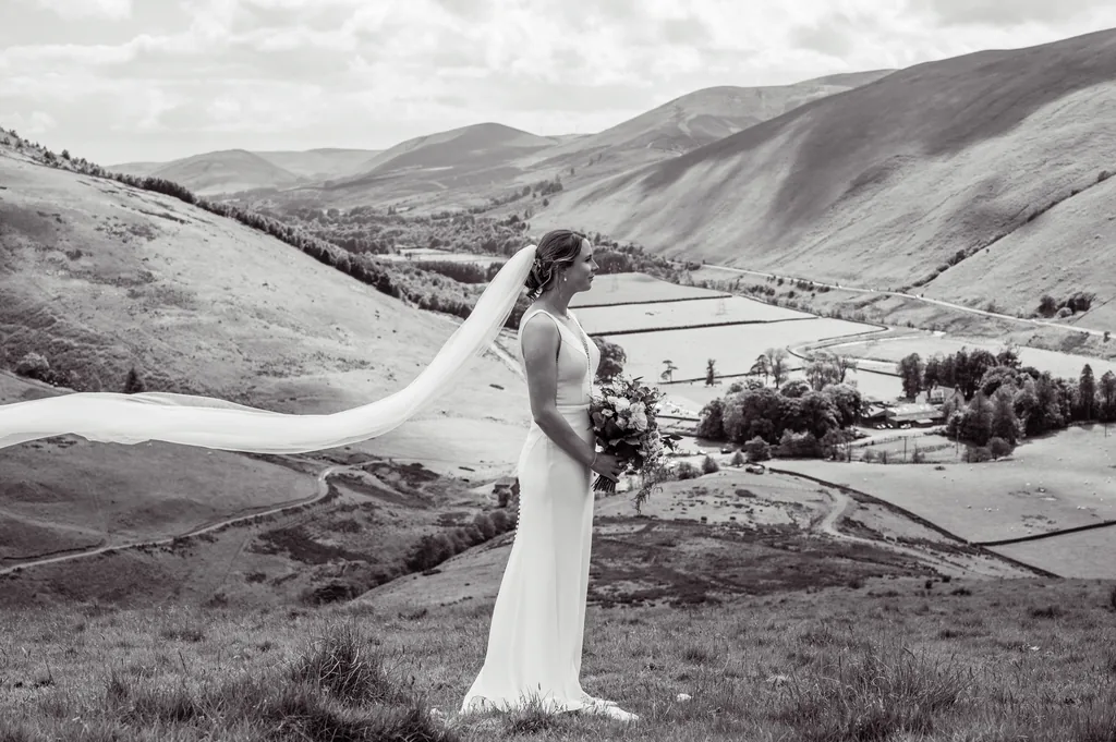 Black-and-white portrait of a bride standing on a hillside overlooking a valley with her veil trailing