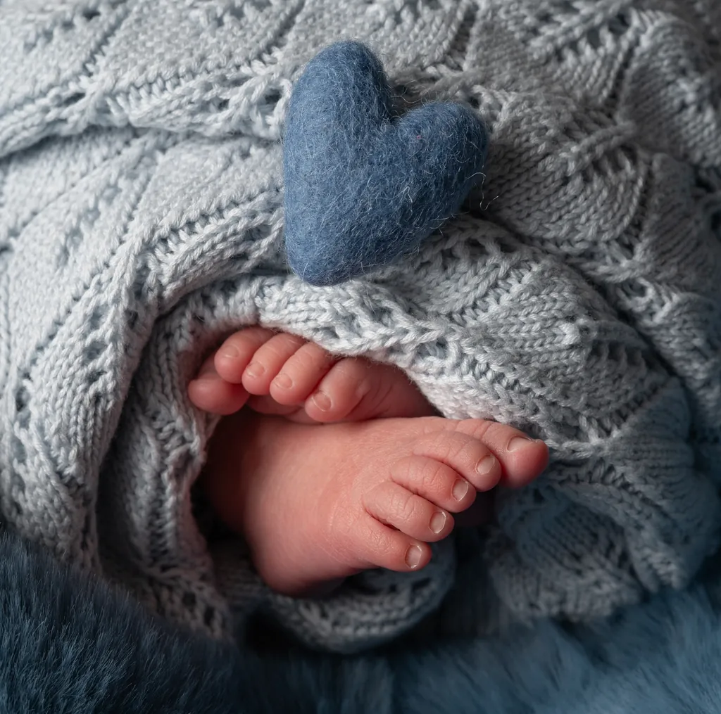 A newborn's tiny feet beside a blue felt heart on a knitted blanket