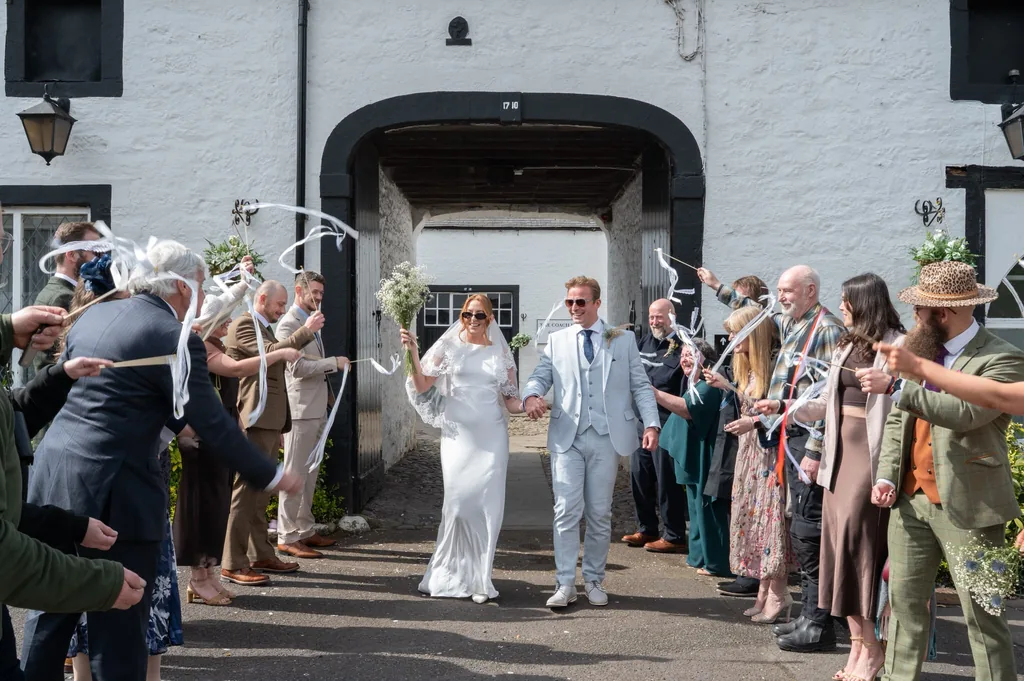 Bride and groom walking out to cheering guests waving ribbon wands outside a white building