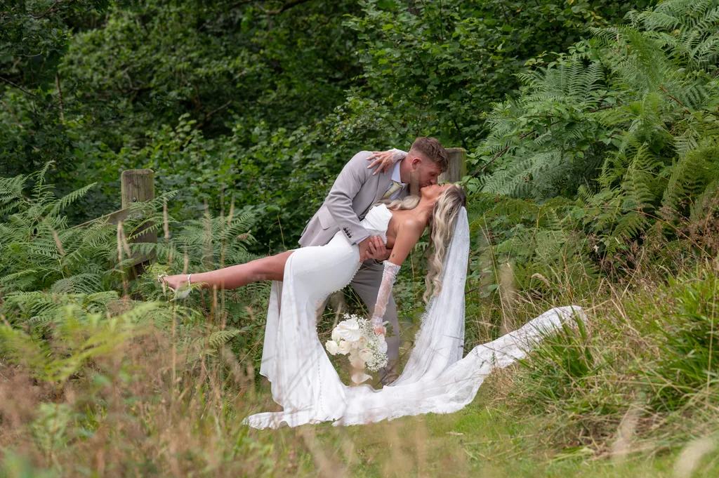 Groom dipping and kissing the bride among ferns in a lush green woodland