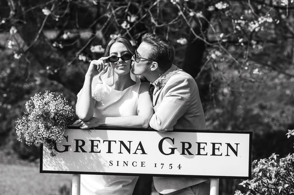Black-and-white photo of a bride and groom behind the “Gretna Green since 1754” sign