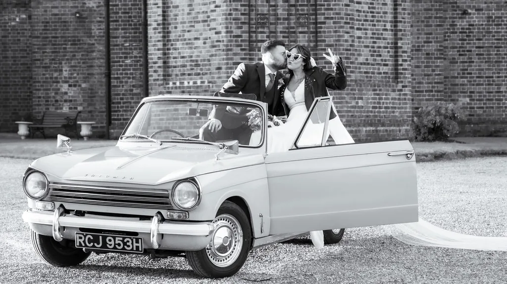 Black-and-white photo of a bride and groom in a white vintage Triumph convertible
