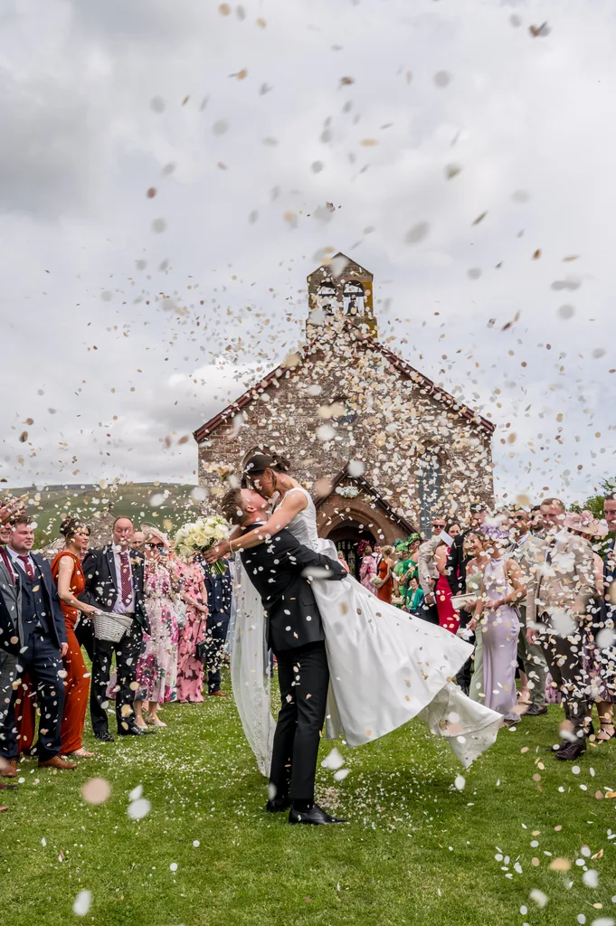 Bride and groom laughing as guests throw a shower of white confetti outside the church