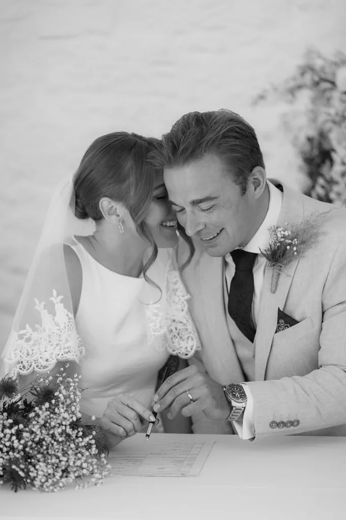 Black-and-white photo of a bride and groom laughing as they sign the wedding register