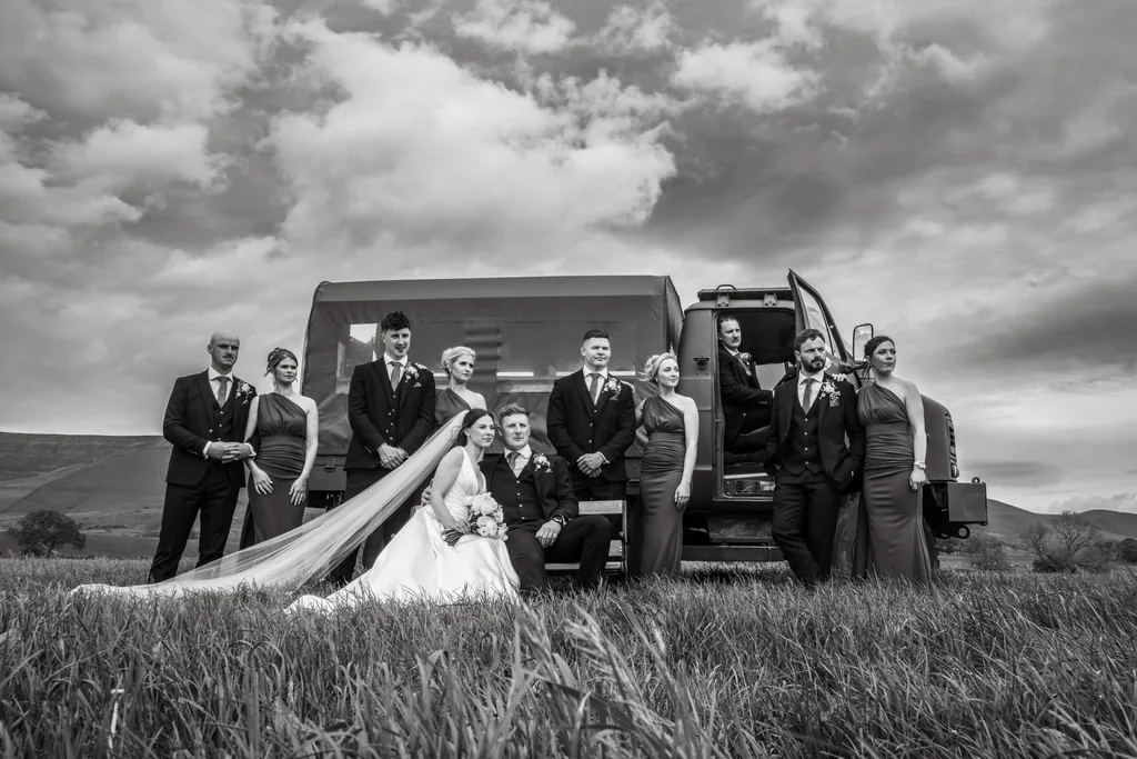Black-and-white portrait of a bridal party celebrating in a field beside a vintage military truck